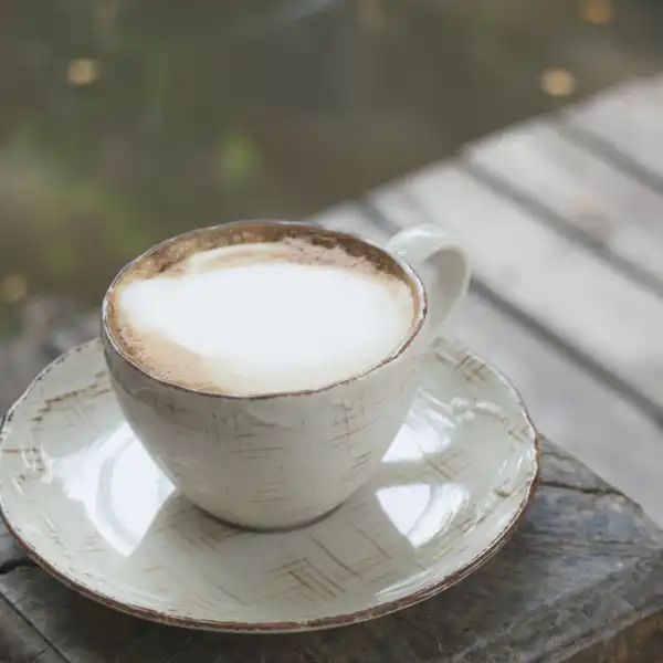 A cream stoneware cup and saucer with a brushed glaze and a brown rim, on a wooden block outdoors