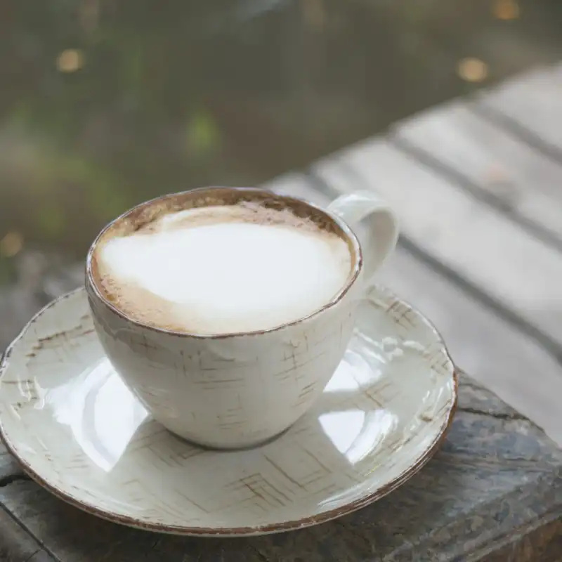 A cream stoneware cup and saucer with a brushed glaze and a brown rim, on a wooden block outdoors