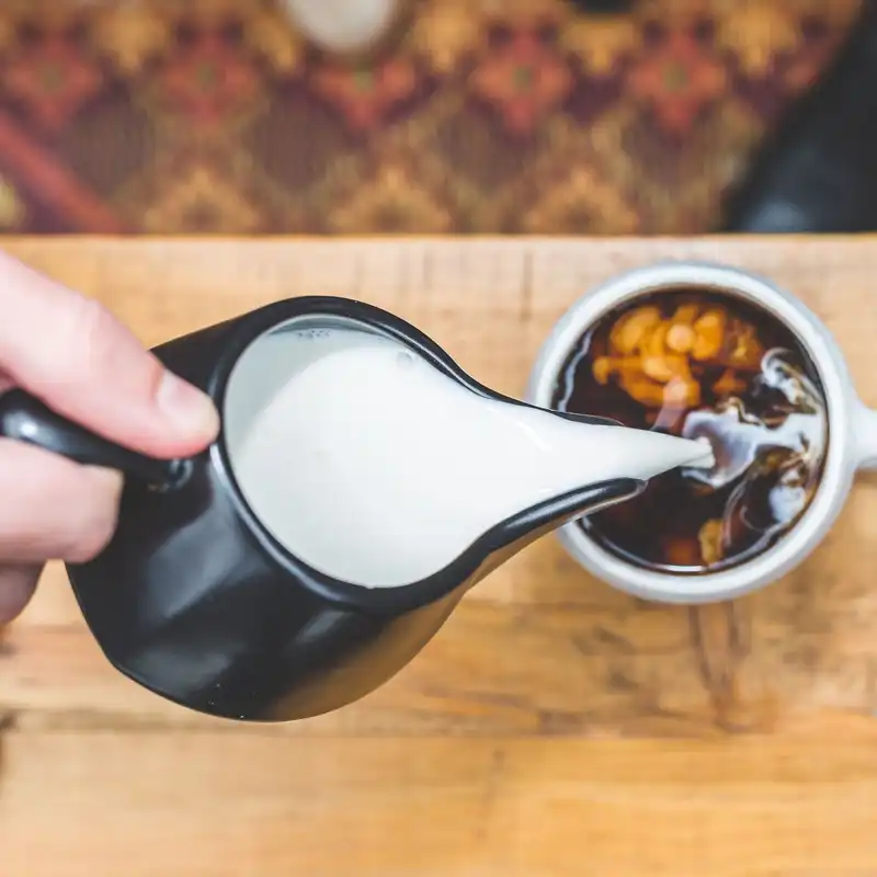 A black glazed jug pouring milk into a cup of coffee on a wooden table