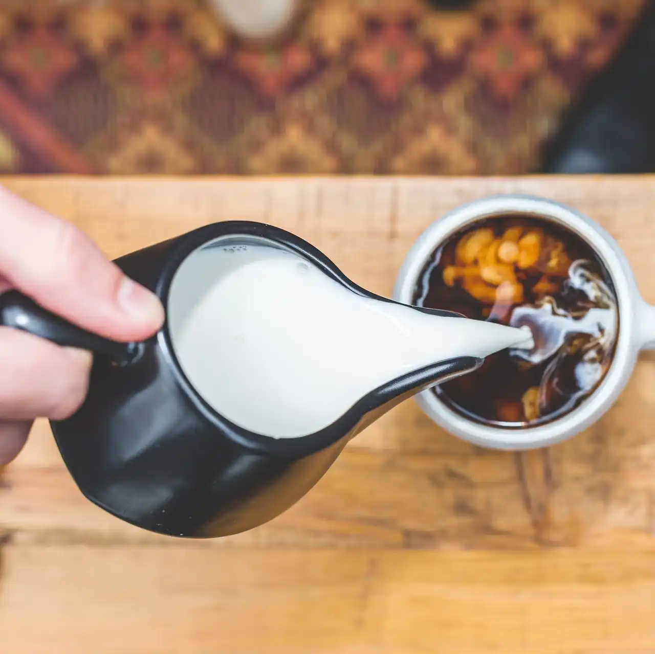 A black glazed jug pouring milk into a cup of coffee on a wooden table