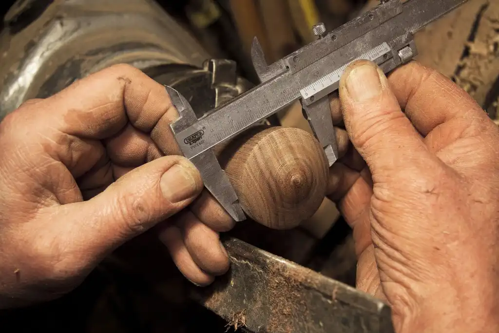 Weathered hands measuring a turned wooden piece with steel calipers