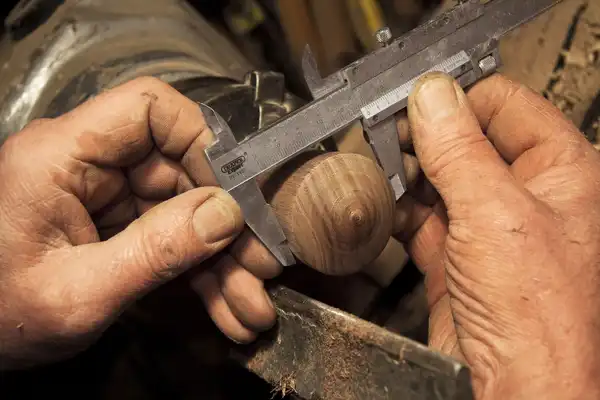 Weathered hands measuring a turned wooden piece with steel calipers