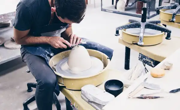 A potter at the wheel shaping a tall white form, tools laid out on the bench beside him