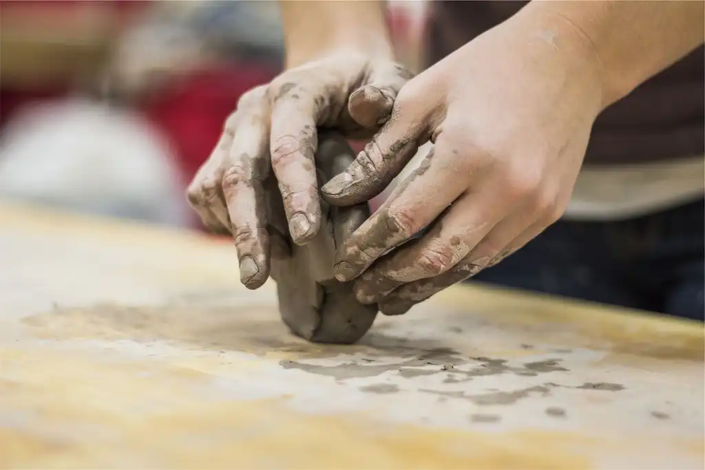 Clay-covered hands kneading a lump of clay on a wooden board