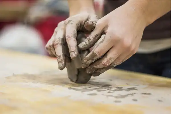 Clay-covered hands kneading a lump of clay on a wooden board
