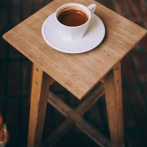 A square-topped wooden stool with crossed stretchers, a cup of coffee on the seat