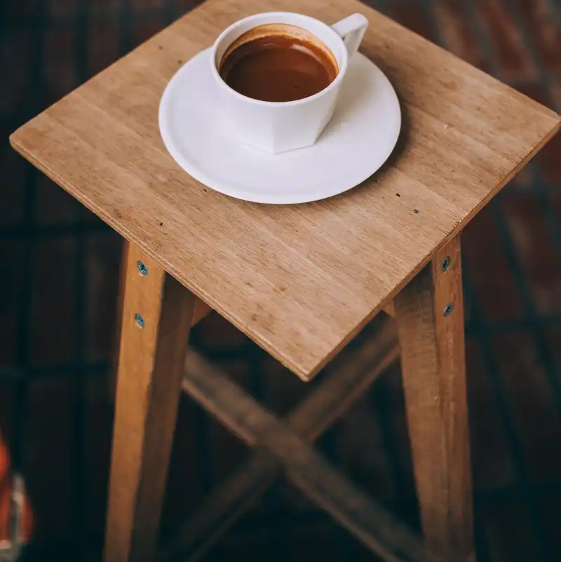 A square-topped wooden stool with crossed stretchers, a cup of coffee on the seat